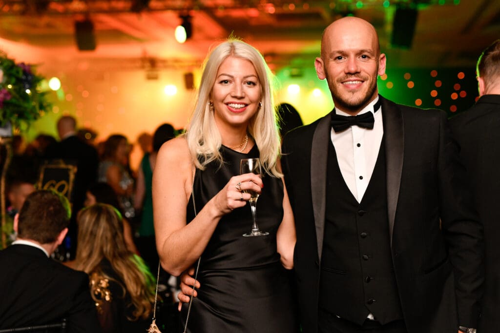 A well lit, elegantly dressed couple poses for the camera with a glass of champagne in hand with green and orange lights fill a ballroom behind them. Great guest shot example to look for when hiring a corporate event photographer.