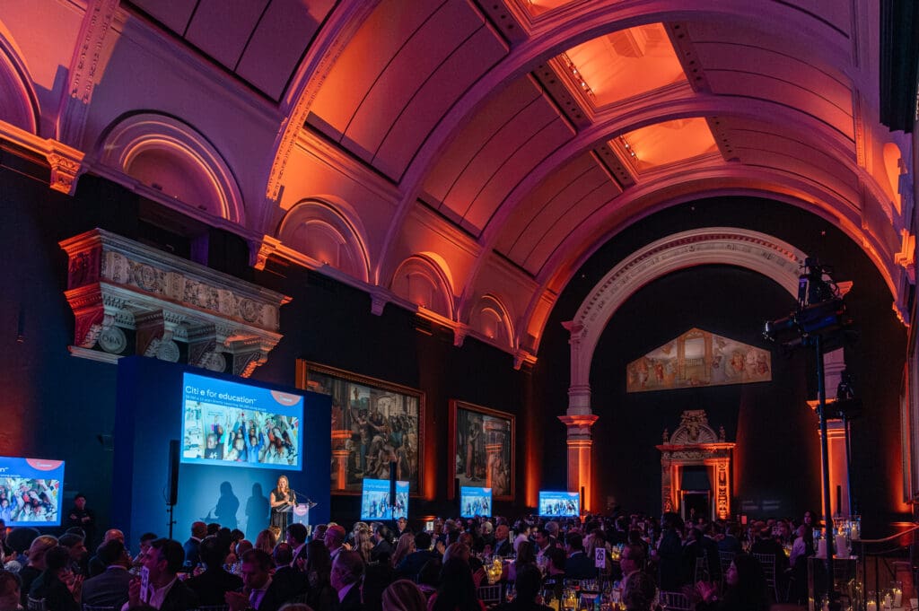 A corporate fundraiser fills the grand ballroom of the Victoria & Albert Museum, with a woman speaking on stage to a large seated audience. Good example of establishing shot, another thing to look for when hiring a corporate event photographer.